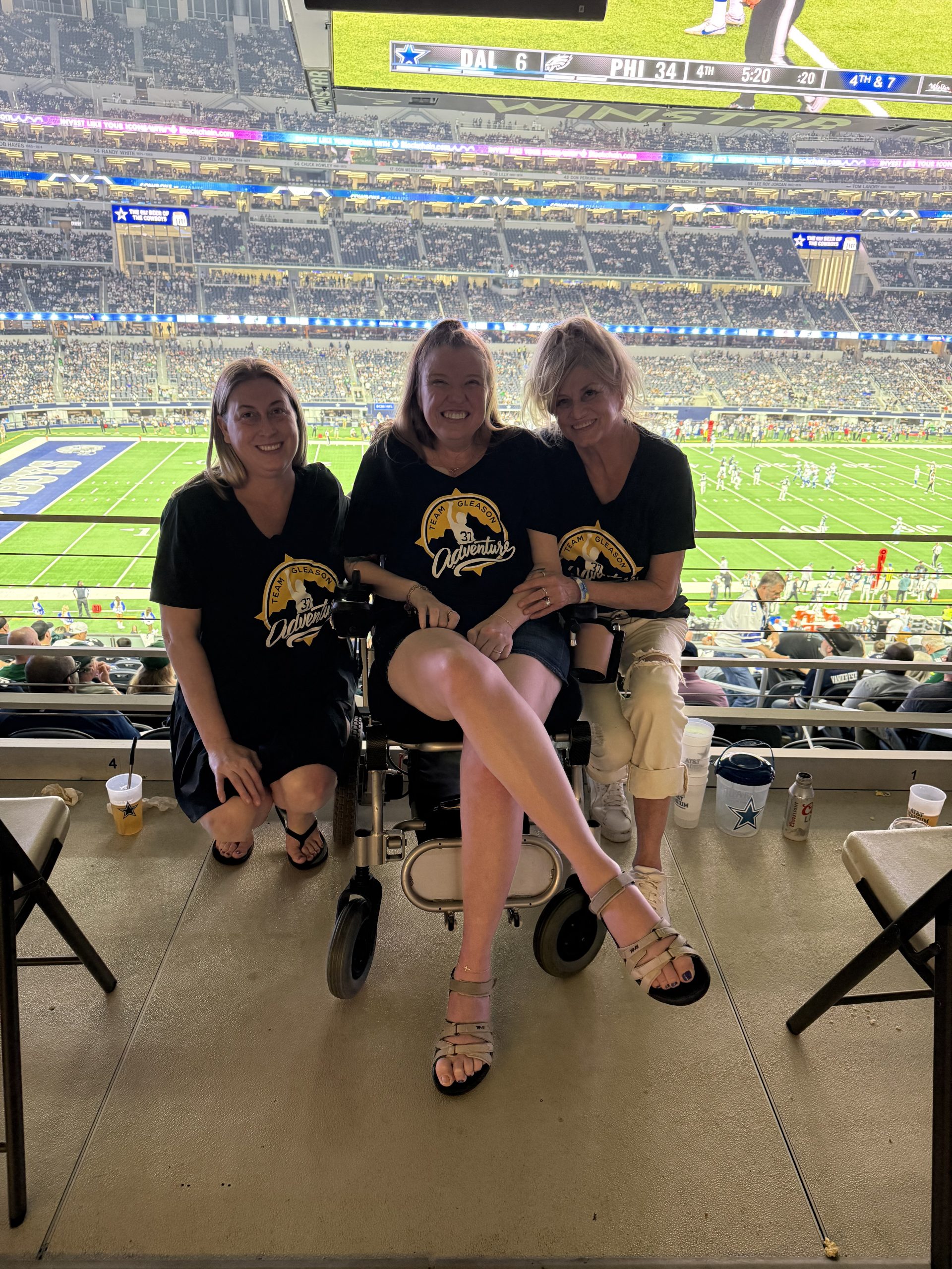 IMG_7428 Sarah Durand Three women are sitting on a balcony with Cowboys stadium field behind them. The woman in the middle is in a wheelchair. The women on either side are kneeling down.