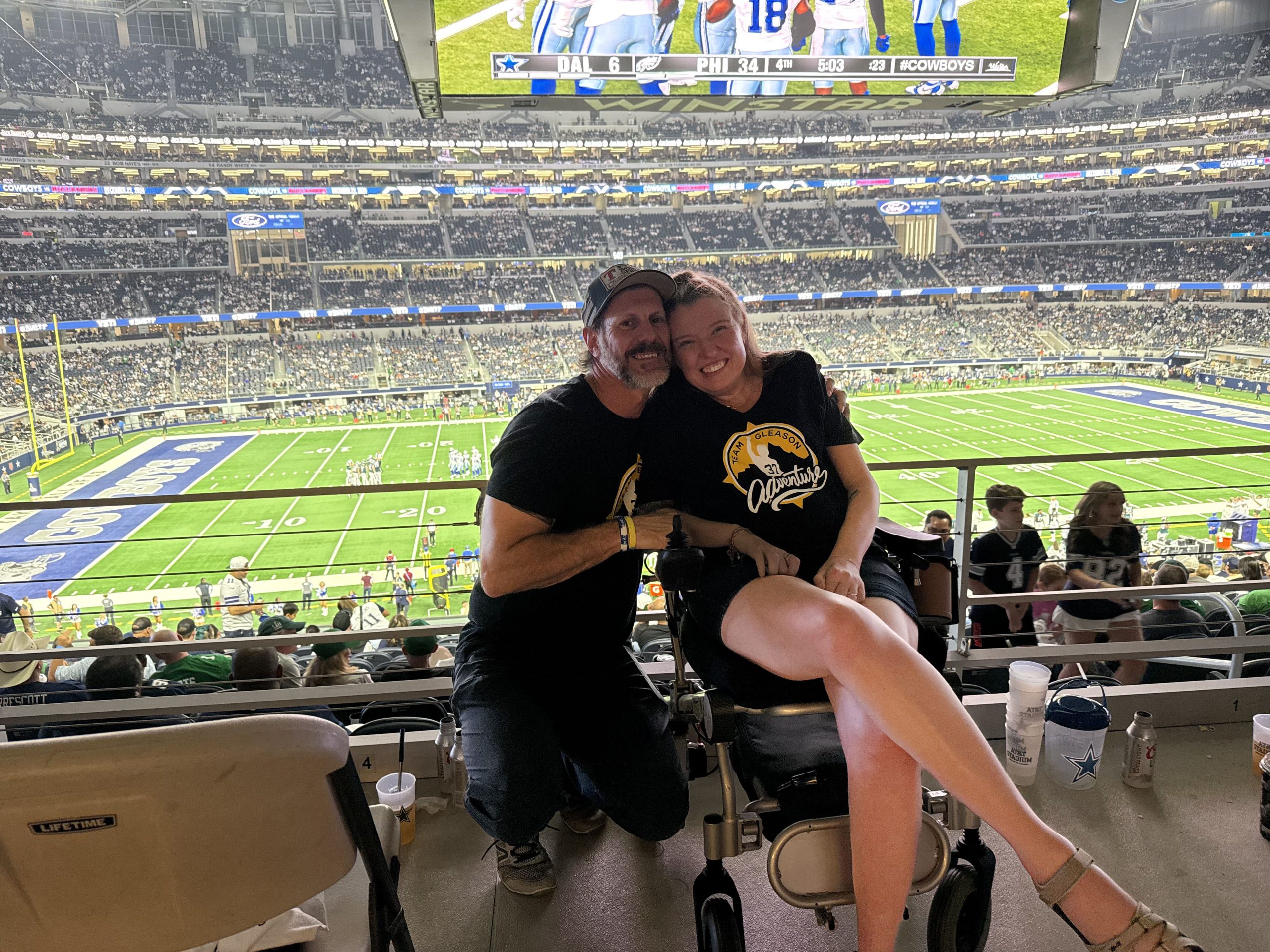 IMG_7436 Sarah Durand edited Two people are on a balcony with the Cowboys stadium field behind them. A woman sits in a wheelchair and a man kneels beside her.